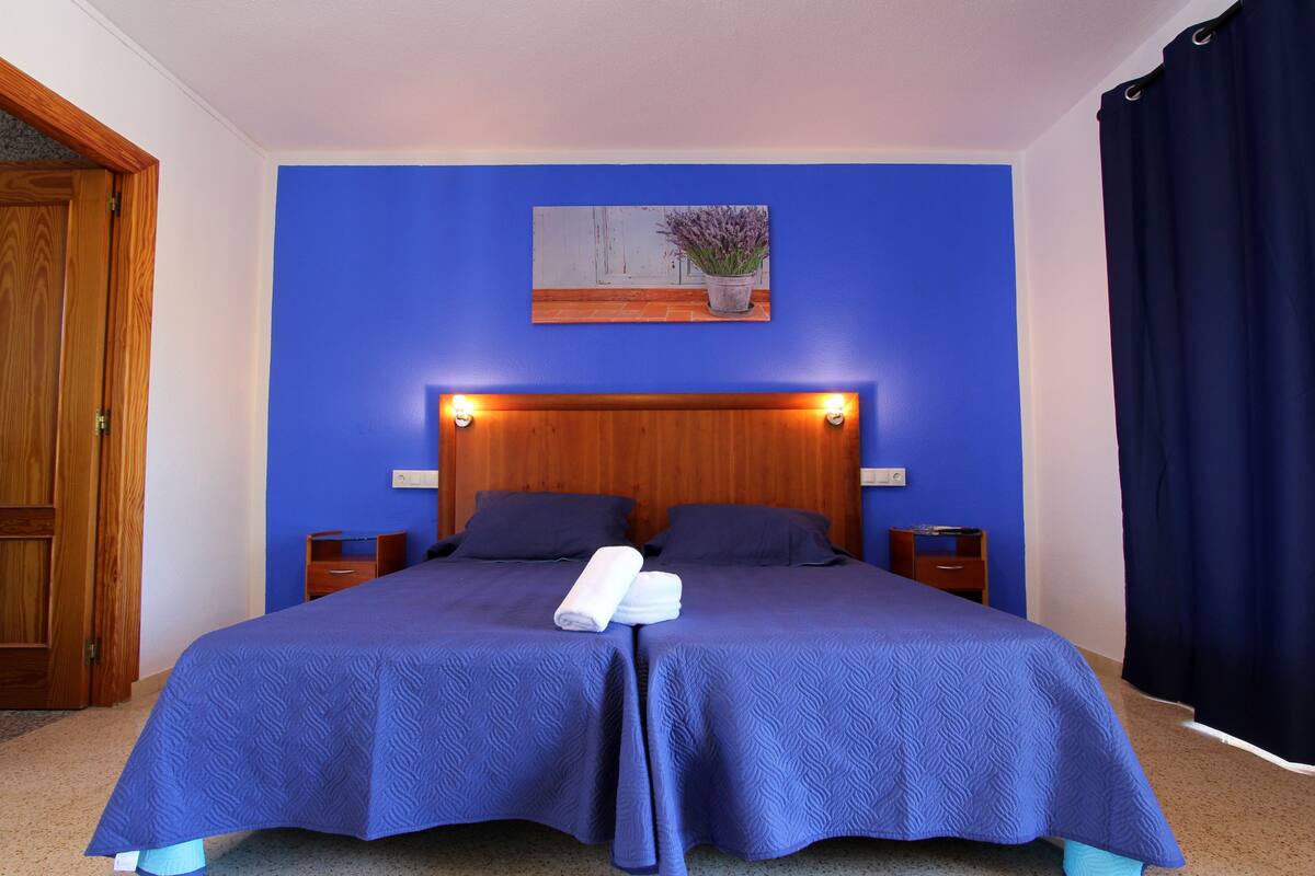 A double bed with a blue bedspread, two pillows, a folded towel, and a nightstand with a lamp.