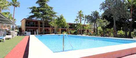 A swimming pool with a red border, surrounded by palm trees and a building with multiple balconies.