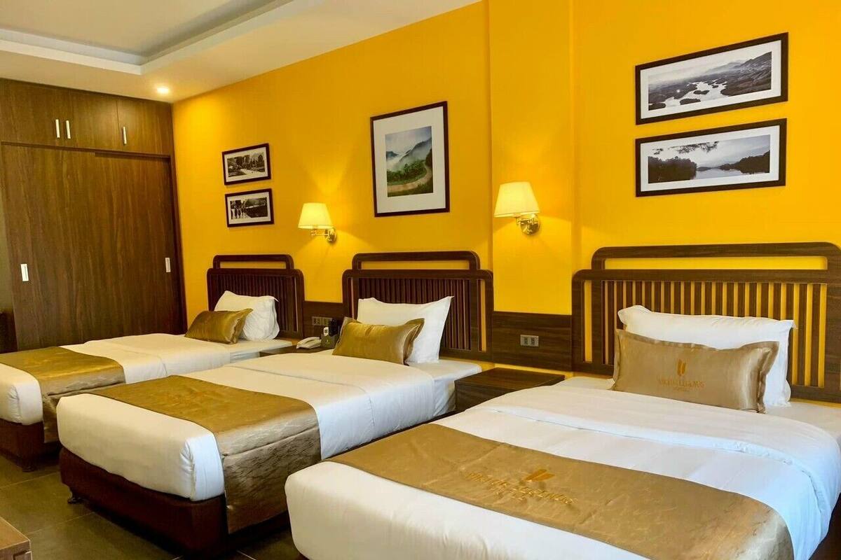 Een hotelkamer met drie enkele bedden, elk met een bruin hoofdbord en witte beddengoed. Twee ingelijste foto’s hangen aan de muur, een houten kledingkast en een nachtkastje met een lamp.