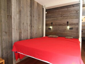 Cama com colcha vermelha em quarto com paredes de madeira e duas luminárias fixadas na parede.