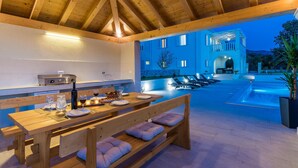 Een terras met een houten tafel en banken, een ingebouwde barbecue en een loungebed bij het zwembad met ligbedden.
