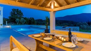 Een terras met eettafel, een zwembad, een houten pergola en uitzicht op een bergachtig landschap.