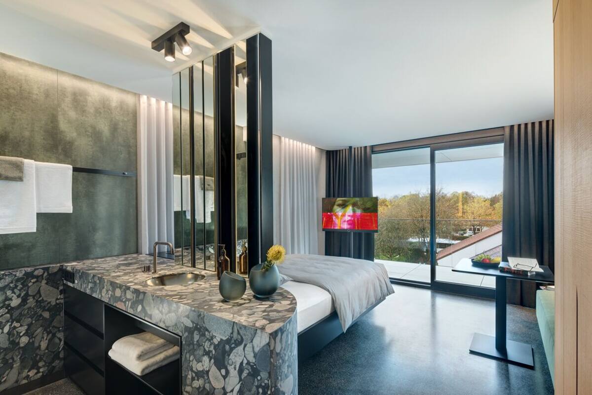 A modern hotel room with a marble bathroom, a large bed, a TV, and a balcony with a city view.