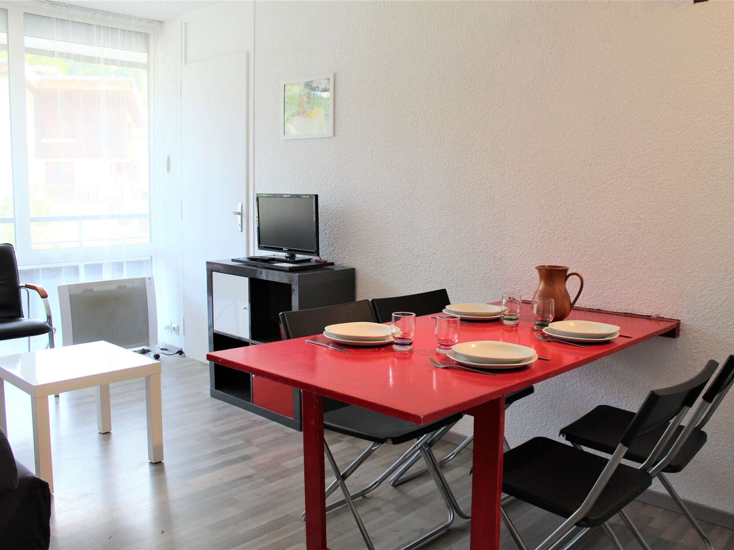 A dining area with a red table, black chairs, and a television on a cabinet.