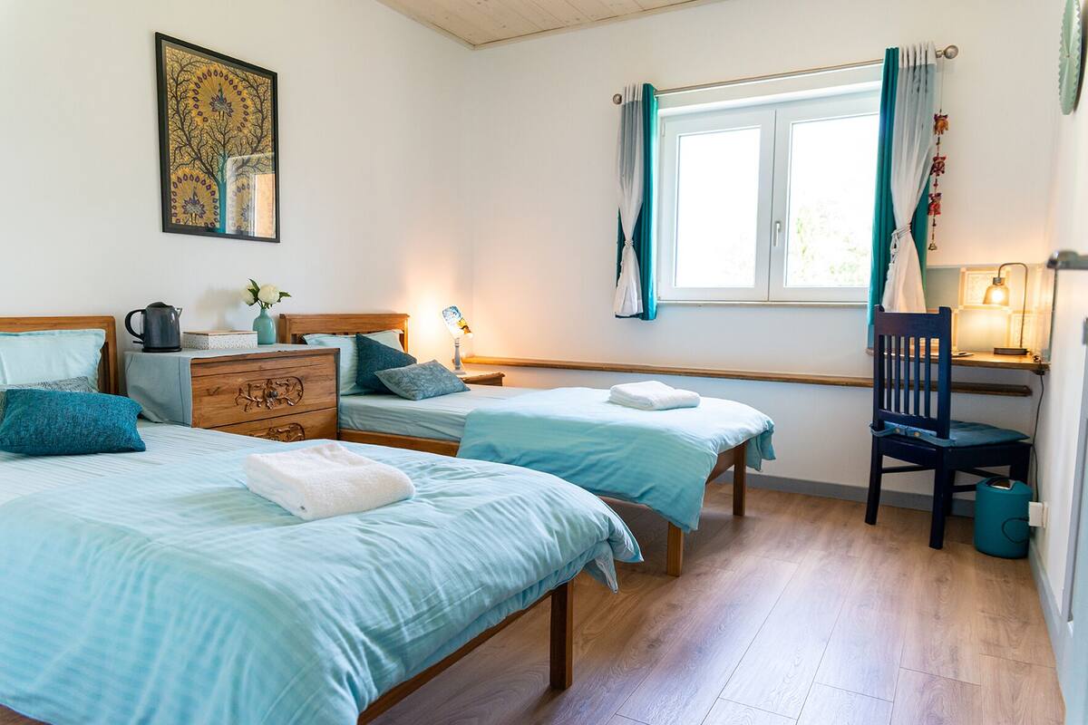 Slaapkamer met twee bedden, een houten dressoir, een bureau met stoel, een raam met gordijnen en een wandlamp.