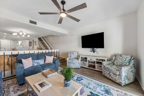 A modern living room with a ceiling fan, a flat-screen TV, a sofa, a patterned armchair, and a coffee table with a plant.
