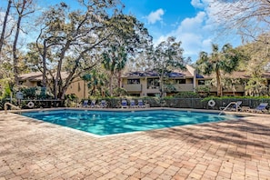 A swimming pool with lounge chairs, surrounded by trees and a building with multiple balconies.