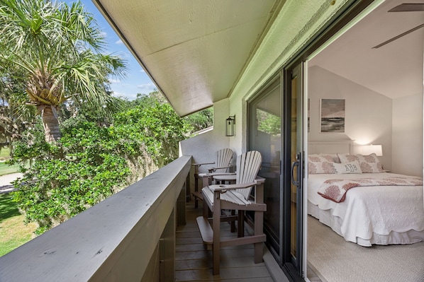 A balcony with a table and chairs, a bed with a white bedspread, and a view of greenery and trees.
