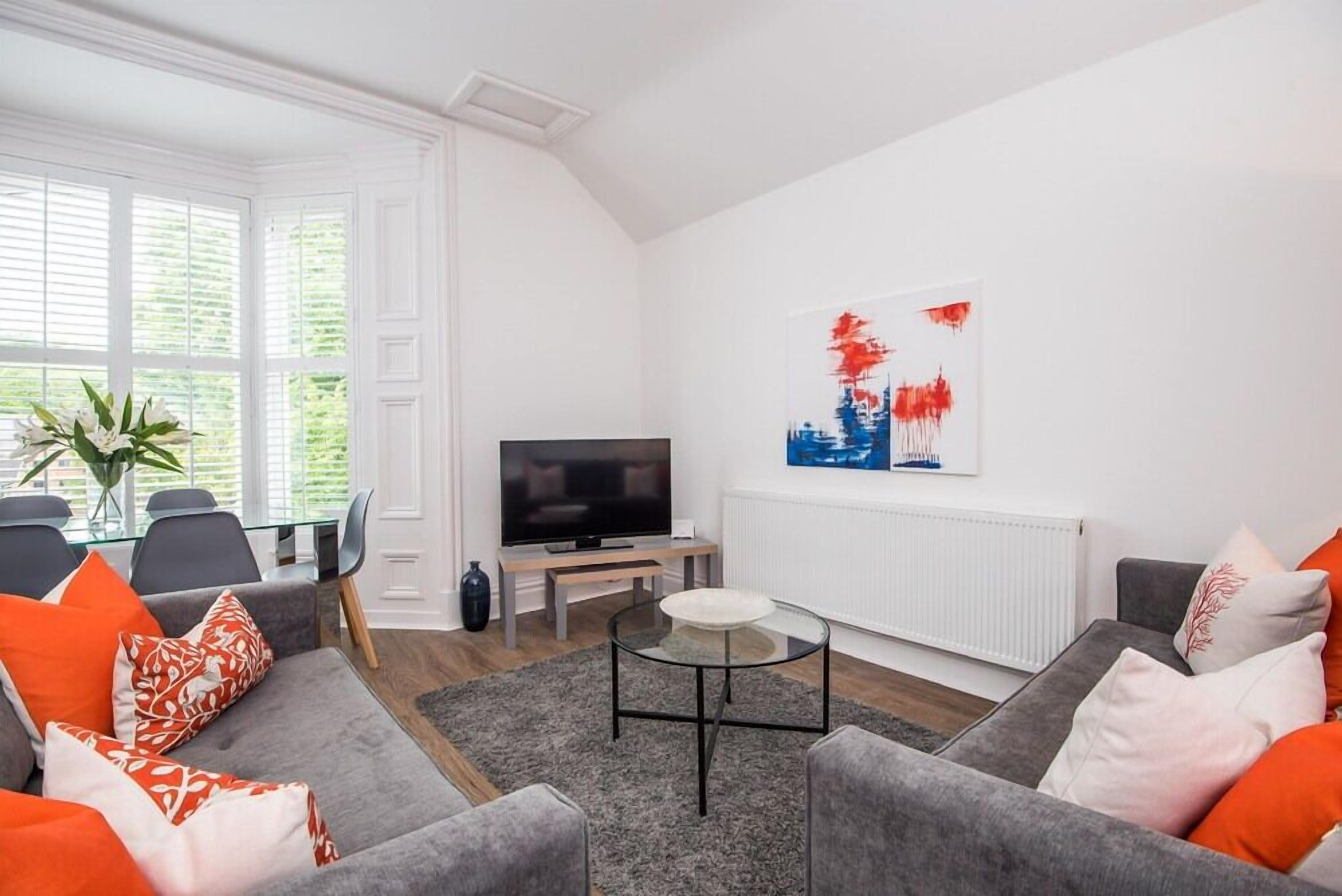 Uma sala de estar moderna com um sofá cinza, uma mesa de centro de vidro, uma televisão de tela plana e uma pintura com vista para a cidade.