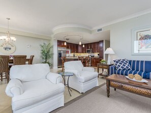 Una sala moderna con un sofá blanco, un sofá azul a rayas y una mesa de centro. Hay una zona de comedor con muebles de mimbre y una cocina al fondo.