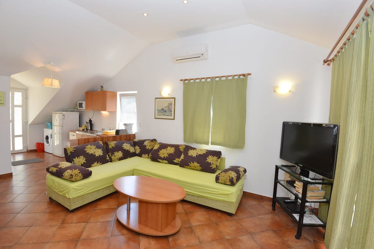 A living room with a sectional sofa, a coffee table, a flat-screen TV, and a kitchen area in the background.