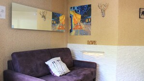 A purple sofa with a cushion, a wooden side table, a wall-mounted mirror, two paintings, a mounted deer head, and a white brick wall with the word "LOVE" on it. - Charming Studio Cabin, Sleeps 4, Central Vars - Next to Ski Lifts (Vars)