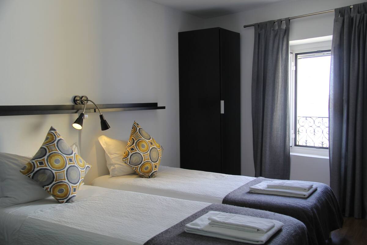 A hotel room with two beds, a large wardrobe, a window with curtains, and a bedside lamp.