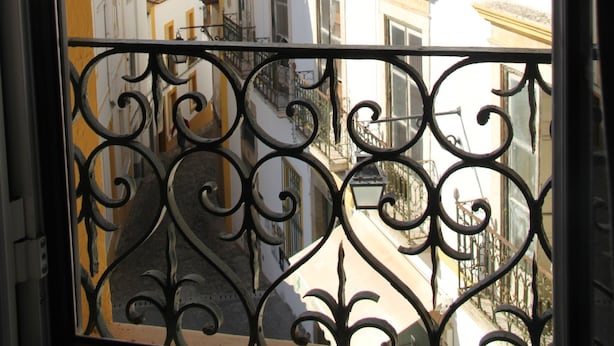 A view through a window with ornate iron railings, featuring a street with buildings that have yellow trim and white facades. - StayInn City - Évora (Évora)
