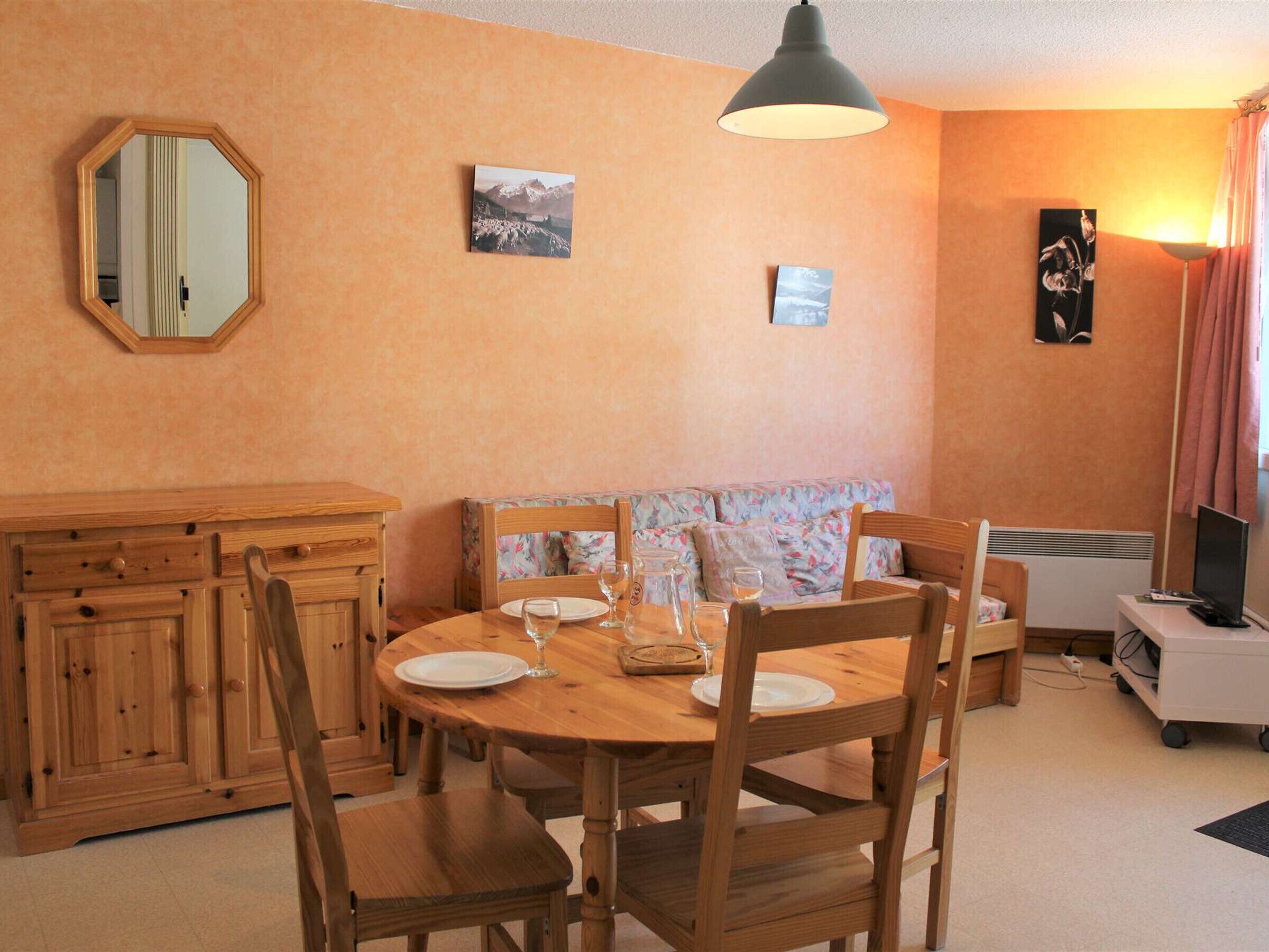 A dining area with a round wooden table, six chairs, a wooden cabinet, a sofa with floral cushions, and a wall-mounted mirror.