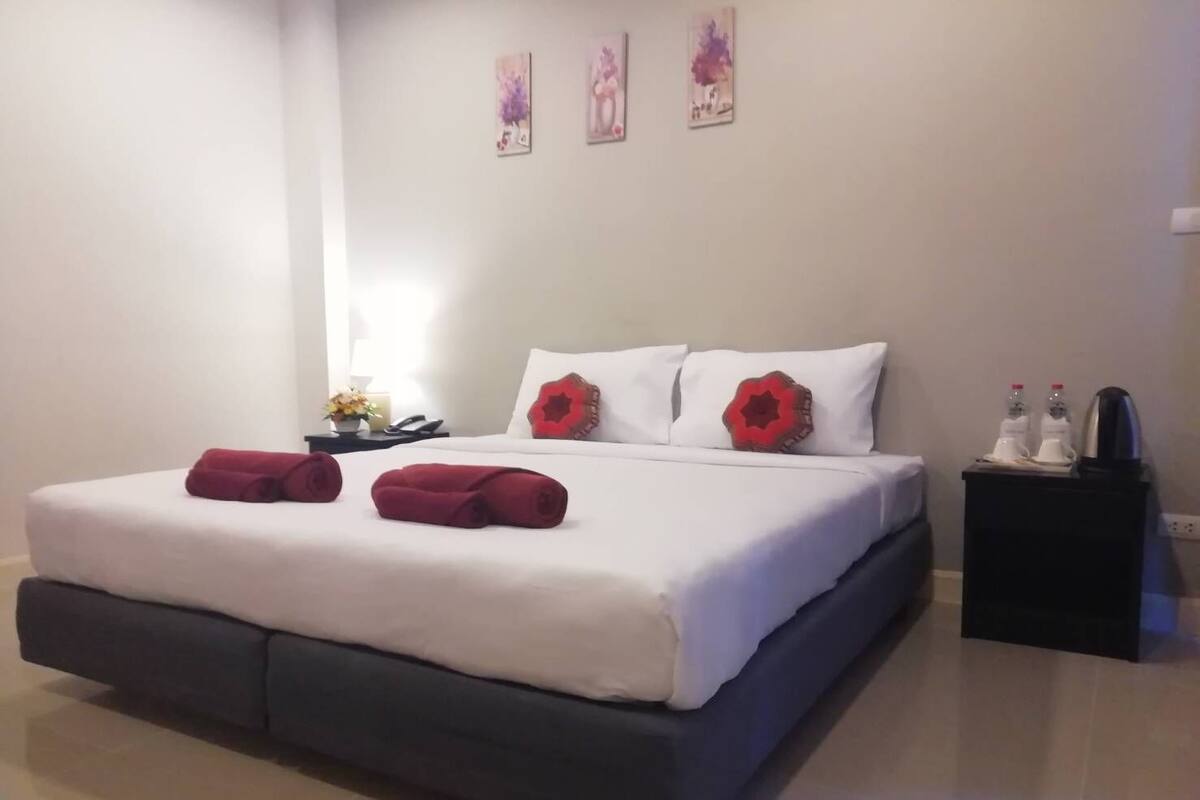 Un lit bien fait, avec du linge de lit blanc et deux coussins décoratifs rouges, une table de chevet avec une lampe et deux bouteilles, et trois œuvres d’art accrochées au mur.