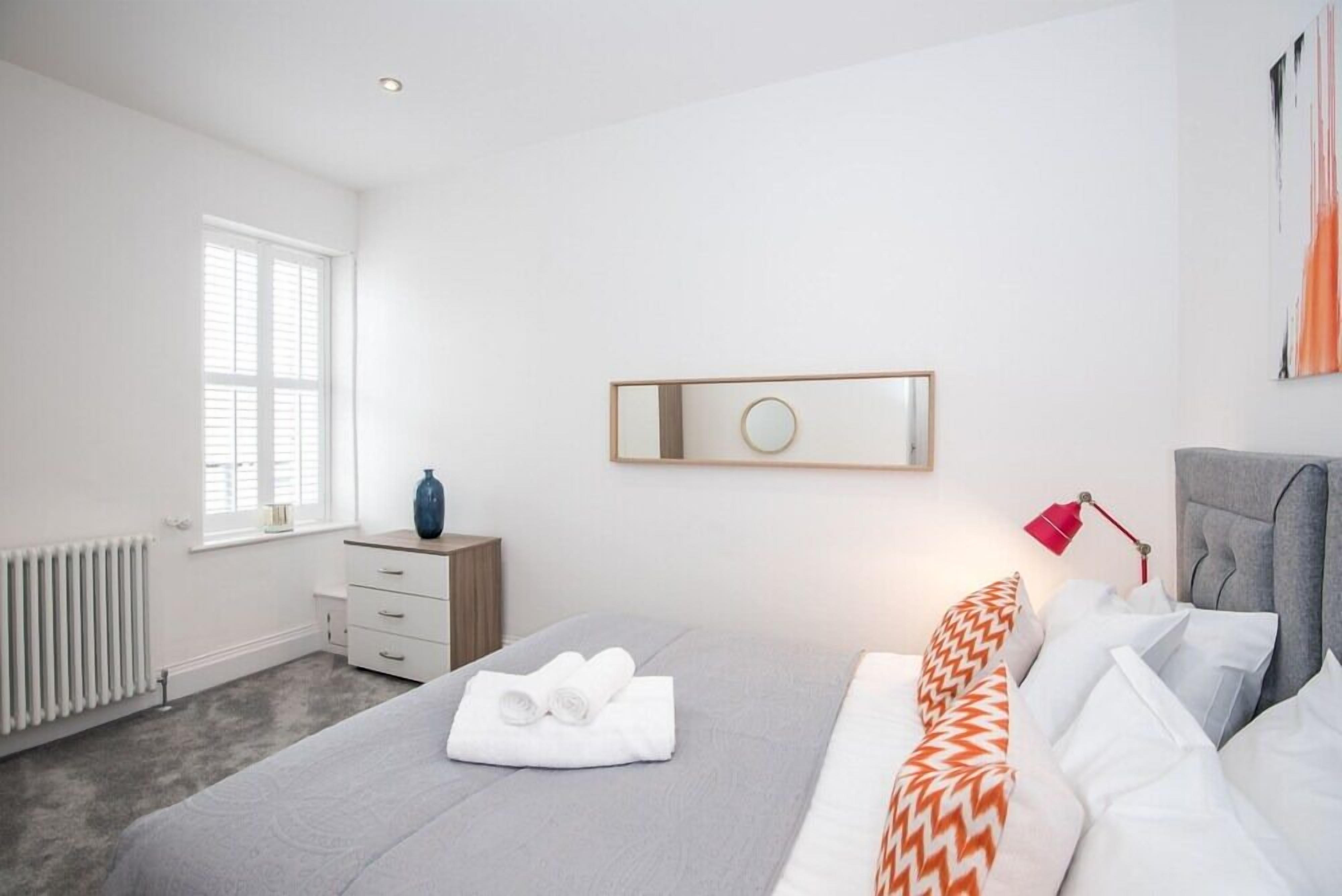 Une chambre moderne avec un lit gris, des coussins blancs et orange à motifs, une table de chevet avec une lampe et une fenêtre avec des stores.
