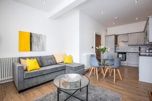 Ein modernes Wohnzimmer mit grauem Sofa, einem Glastisch und einem Essbereich mit kleinem Tisch und Stühlen.