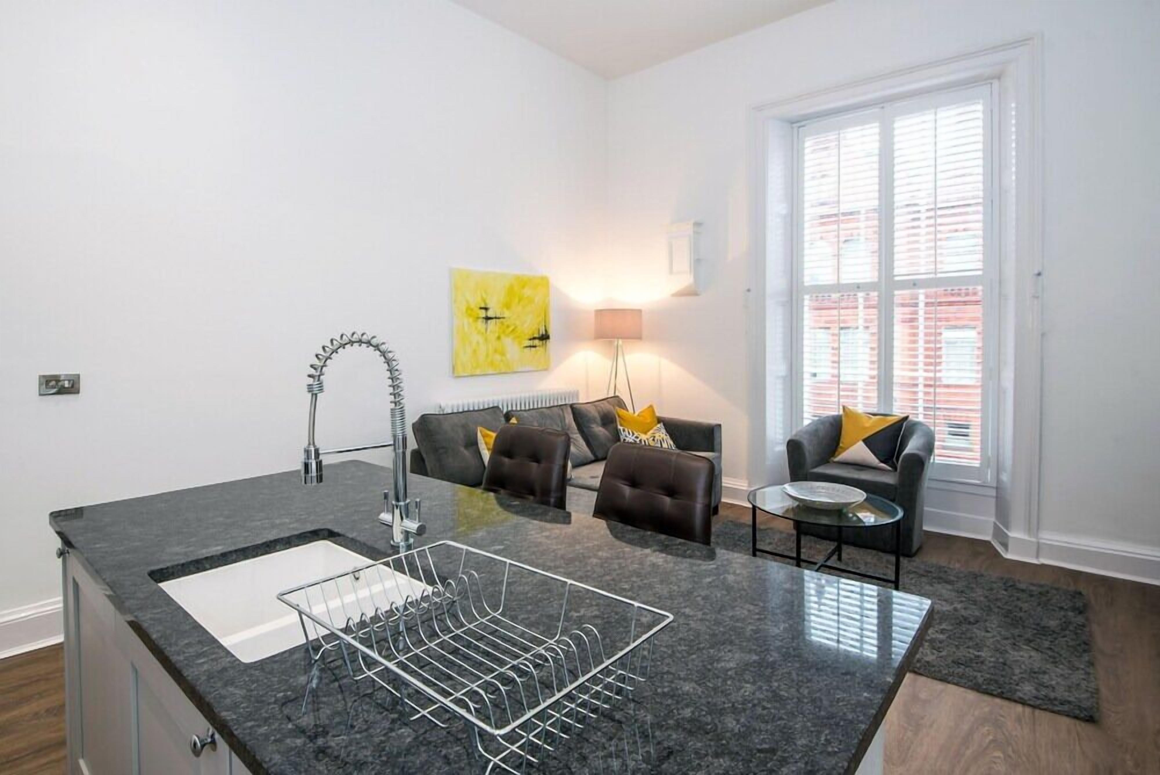 A modern kitchen with a granite countertop, a built-in sink, and a dish rack. There is a seating area with a sofa and armchairs, a small round table, and a lamp. A large window provides natural light.