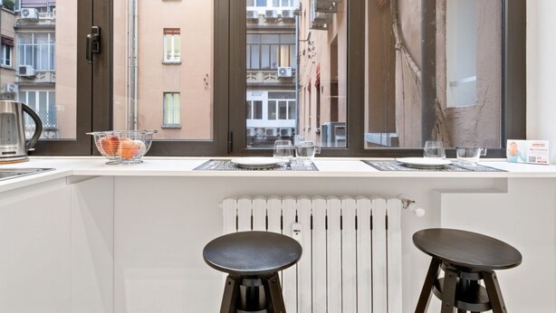Una cocina con ventana que ofrece vistas a un paisaje urbano, una tetera y dos taburetes.