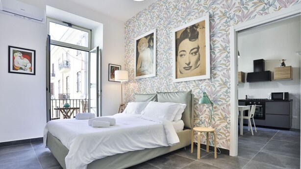 A modern bedroom with a bed, a small table, and a chair. There is a window, a door leading to a kitchen, and a wall with floral wallpaper. - GuestHost - Garibaldi huge and cozy apartment (Napoli)