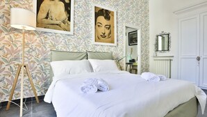 A neatly made bed with white linens and towels, a floor lamp, and a wall with floral wallpaper and framed artwork. - GuestHost - Garibaldi huge and cozy apartment (Napoli)