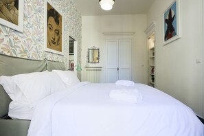 Een net opgemaakt bed met witte lakens, een behang met bloemenmotief en een spiegel erboven.