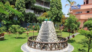 Un jardín con una fuente de piedra rodeada por una cerca de madera, un edificio con un letrero que dice 'Hotel Bedrock', y abundante follaje.