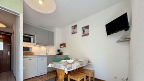 Cozinha compacta com micro-ondas, fogão e máquina de lavar loiça. Há uma mesa de jantar com cadeiras e uma televisão montada na parede.