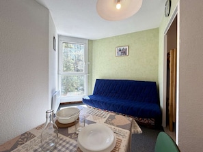 A room with a bed, a table with a bowl and a glass, a window with a view of trees, and a wall clock.