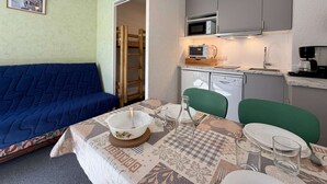 Um espaço compacto com cozinha americana, mesa de jantar para dois, sofá azul e uma cama pequena.