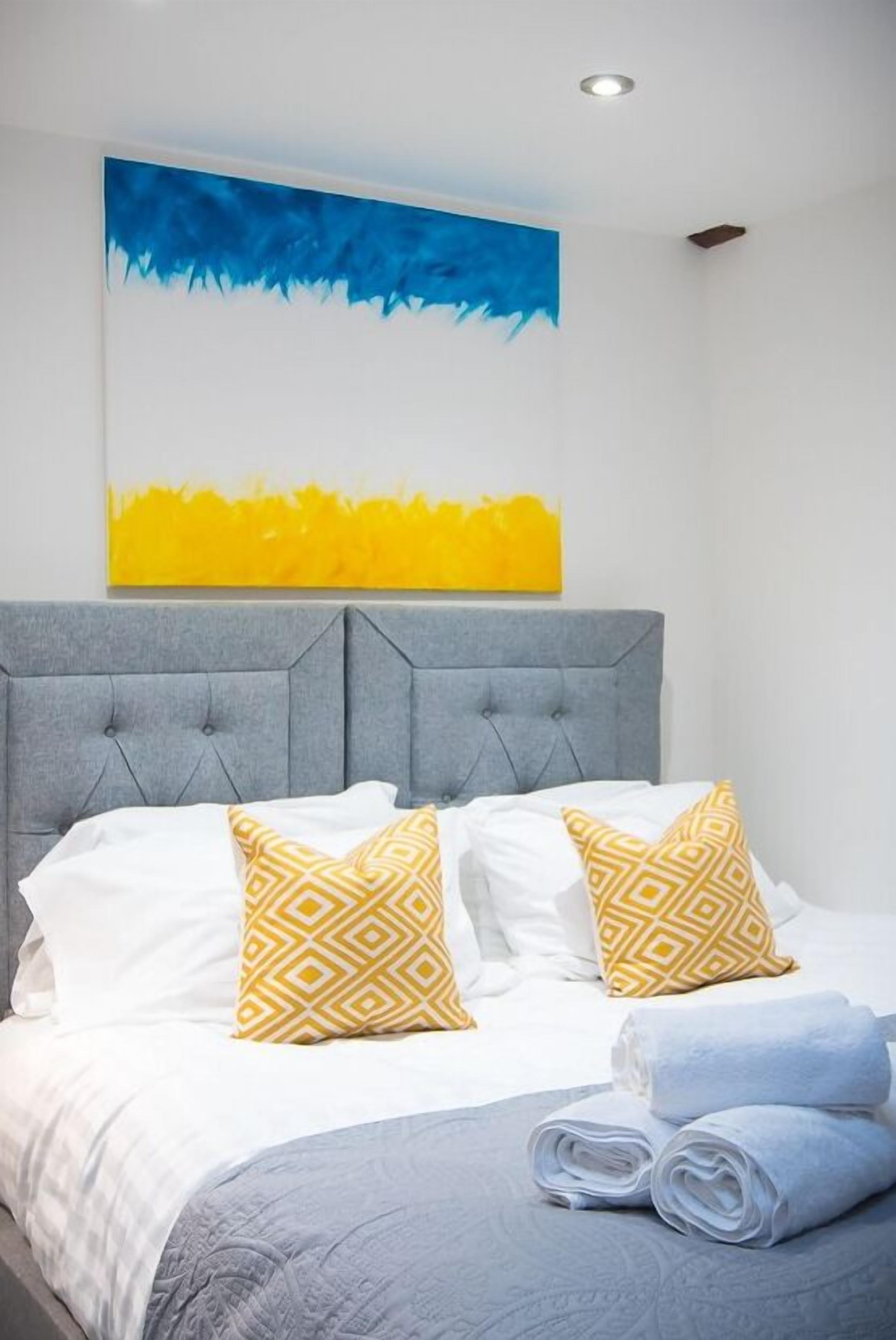 A neatly made bed with white and yellow patterned pillows, a grey headboard, and a blue and yellow abstract painting above.