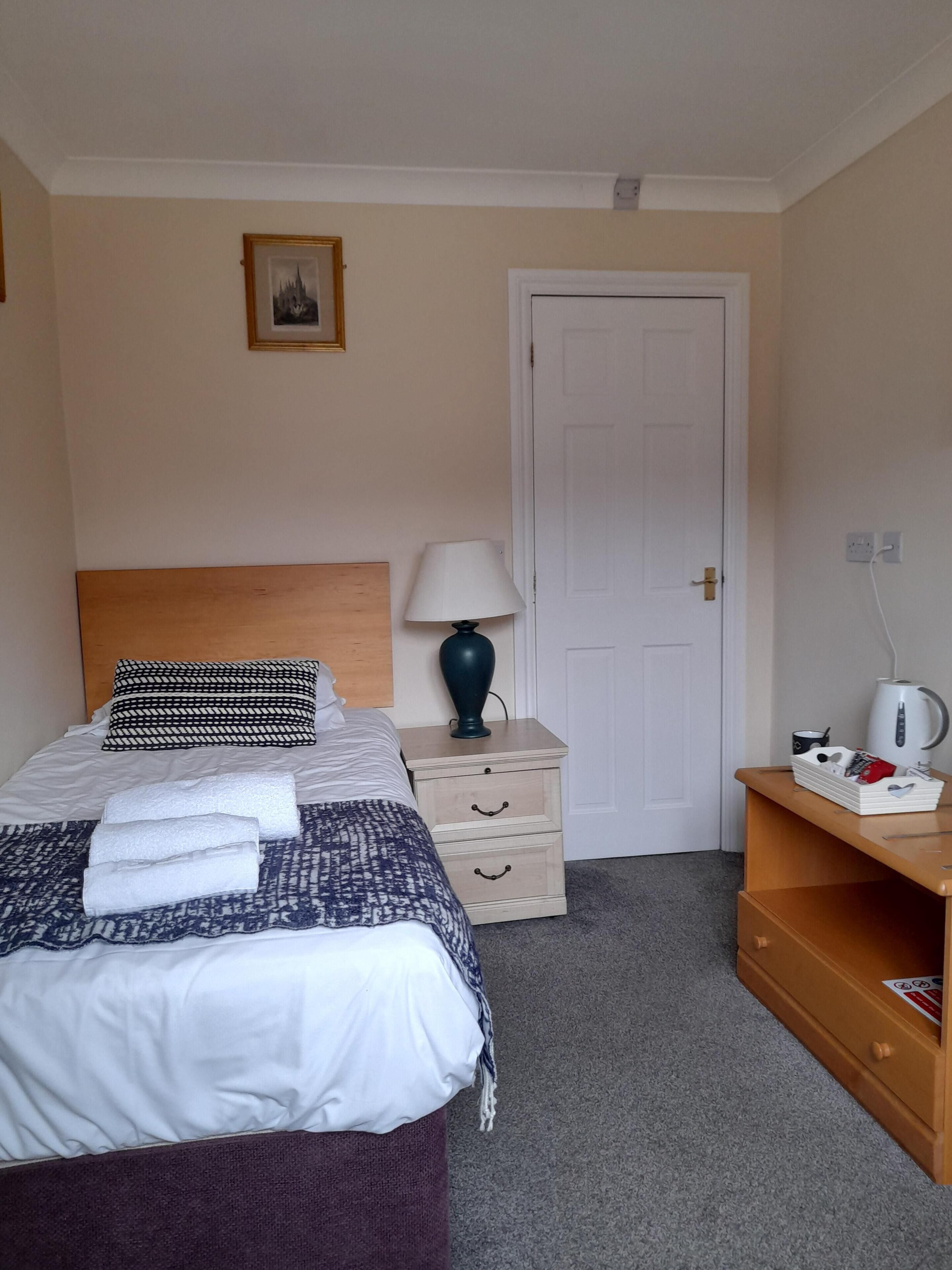 A neatly made bed with a patterned blanket and pillows, a bedside lamp, a small wooden nightstand with drawers, a white door, and a shelf with a kettle and a mug.