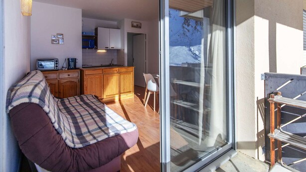 Un salon accueillant avec un canapé, une cuisine dotée de meubles en bois et une porte-fenêtre coulissante donnant sur un balcon avec vue sur une montagne enneigée.
