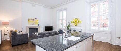 A modern kitchen with a granite countertop, white cabinetry, and a built-in sink. There is a dining area with a gray sofa and a small table.