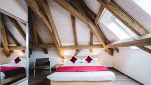 Een slaapkamer met een schuine kap, houten balken, een bed met witte beddengoed en rode accenten, een stoel en een nachtkastje met een lamp.