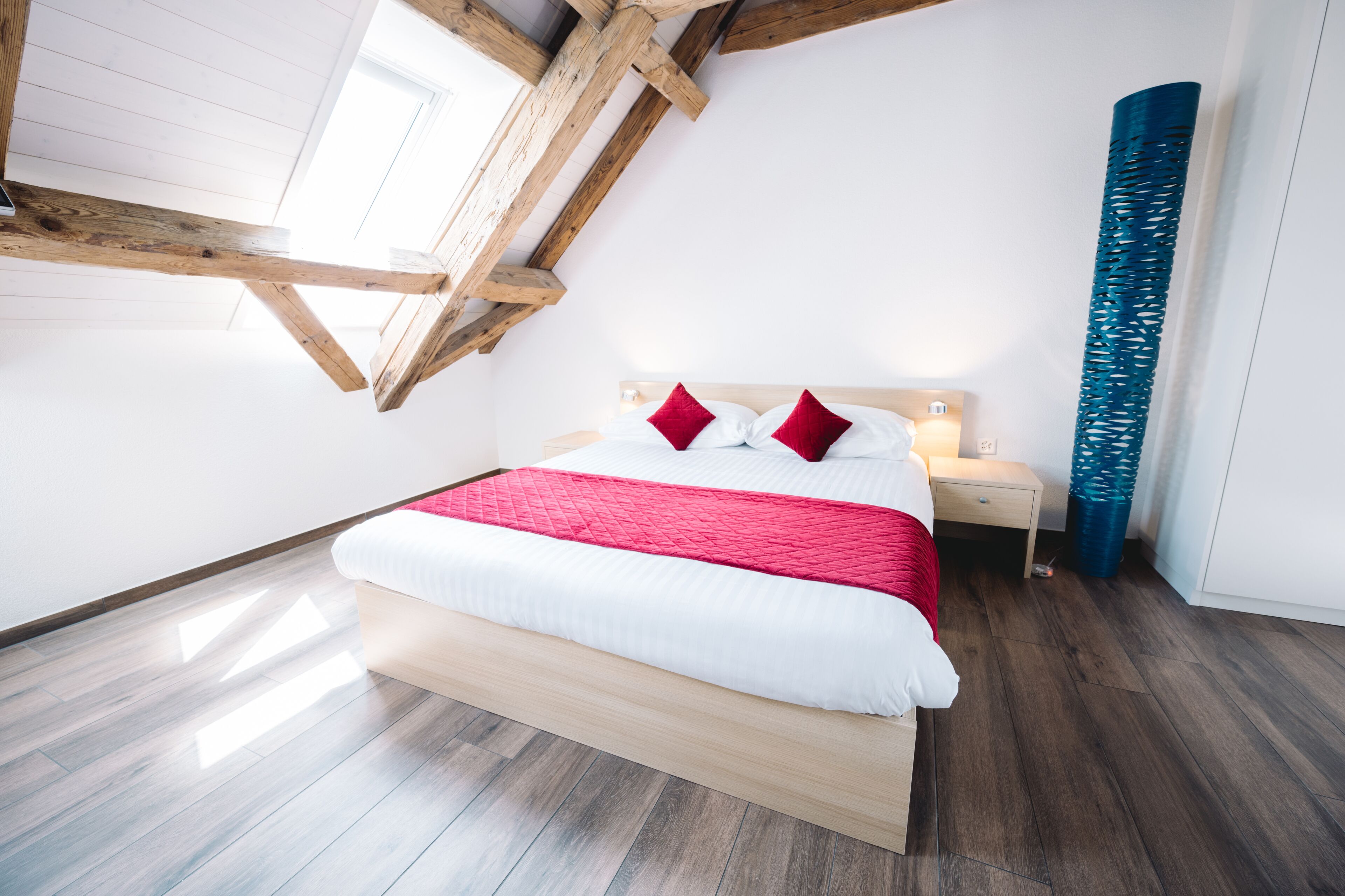 Un dormitorio moderno con techo inclinado, estructura de cama de madera, ropa de cama blanca y una almohada roja de acento.