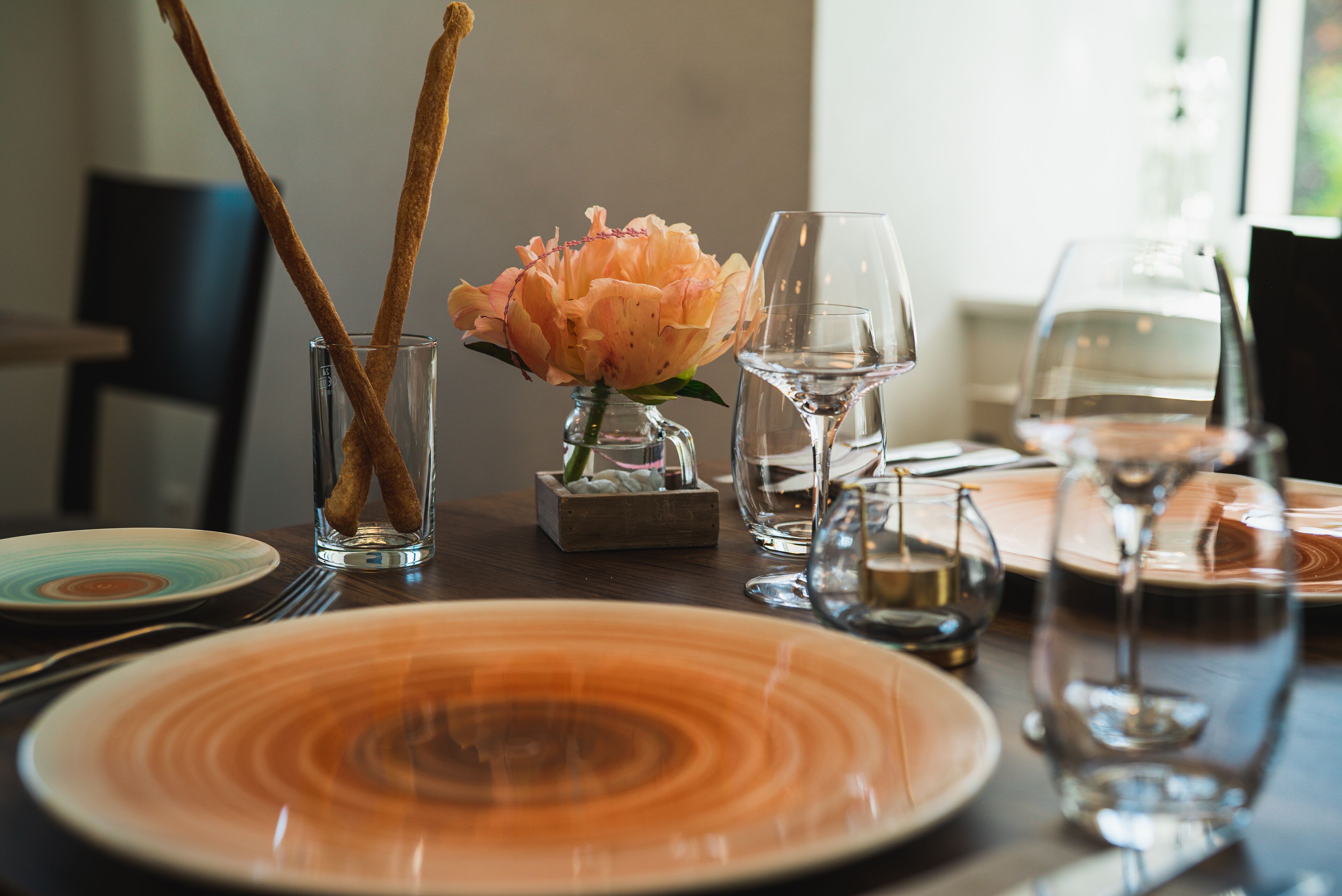 A dining table set with plates, glasses, and a vase with a flower.