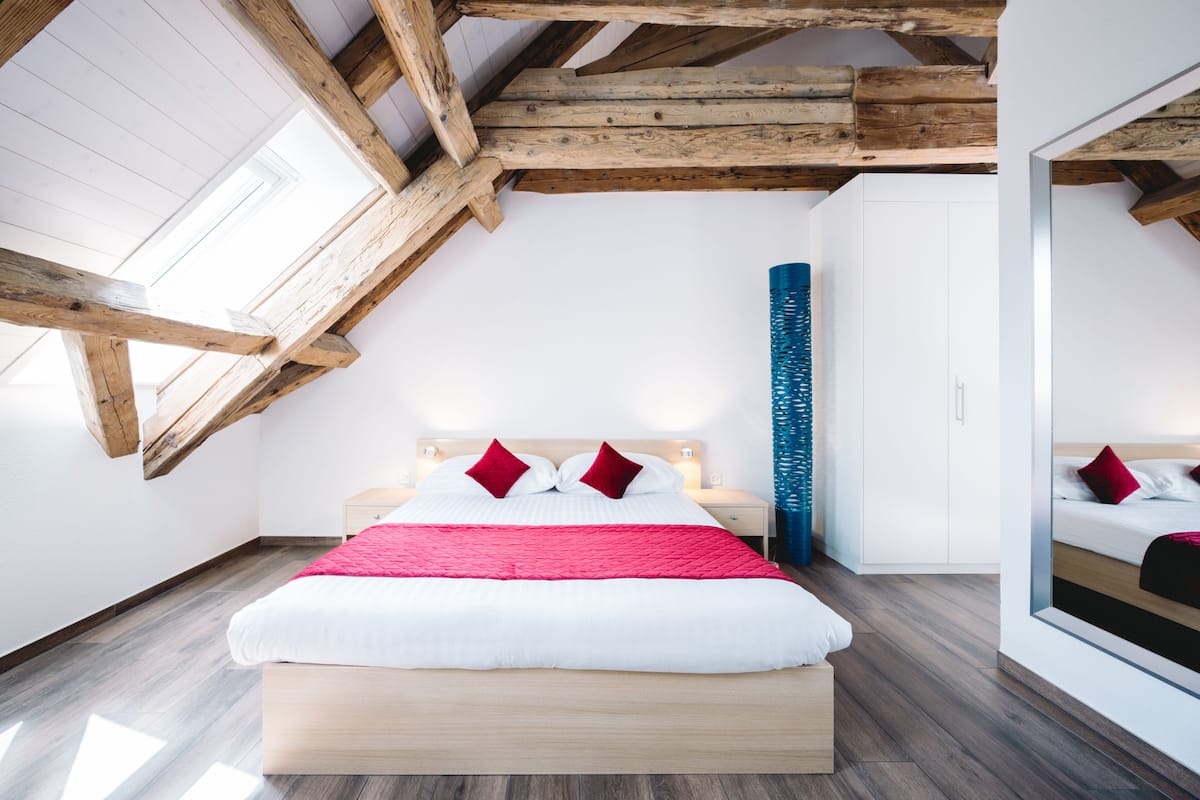 Een moderne slaapkamer met een houten plafond, een groot bed met witte beddengoed en rode kussens, een hoge blauwe vaas en een witte kledingkast.