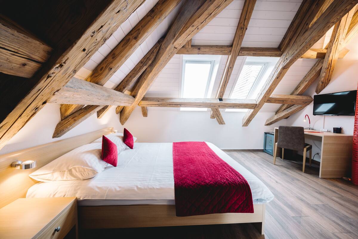 Een slaapkamer met een schuine plafond, een bed met wit beddengoed en rode kussens, een bureau met stoel, en een televisie aan de muur.