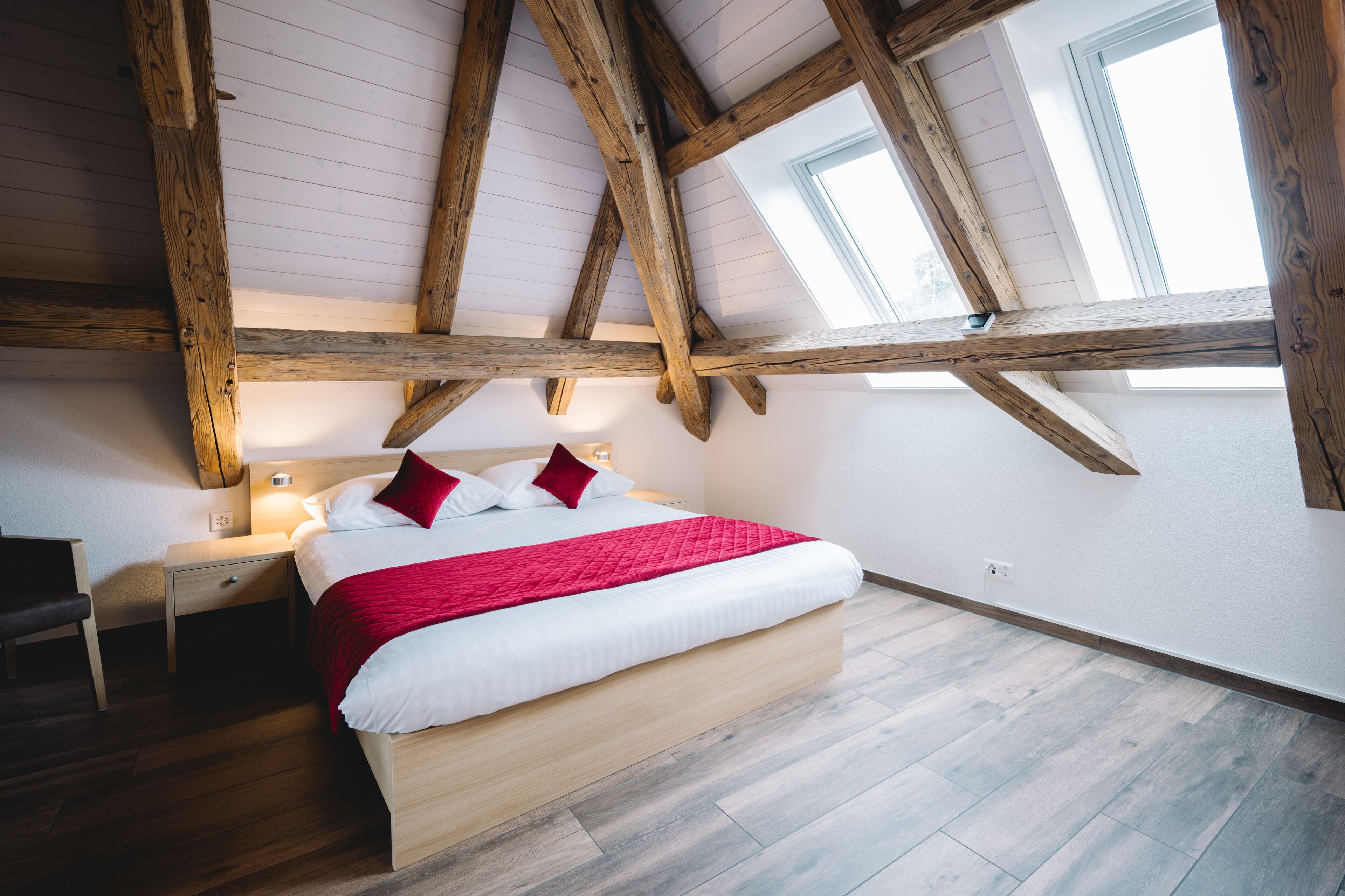 A bedroom with a wooden ceiling, a bed with white bedding and red pillows, a nightstand with a lamp, and a window with a view of the sky.