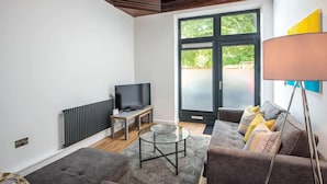 A modern living room with a sofa, a glass coffee table, a TV on a stand, and a large window with a view of greenery. - The Wolsey at 52 Old Elvet (Durham)
