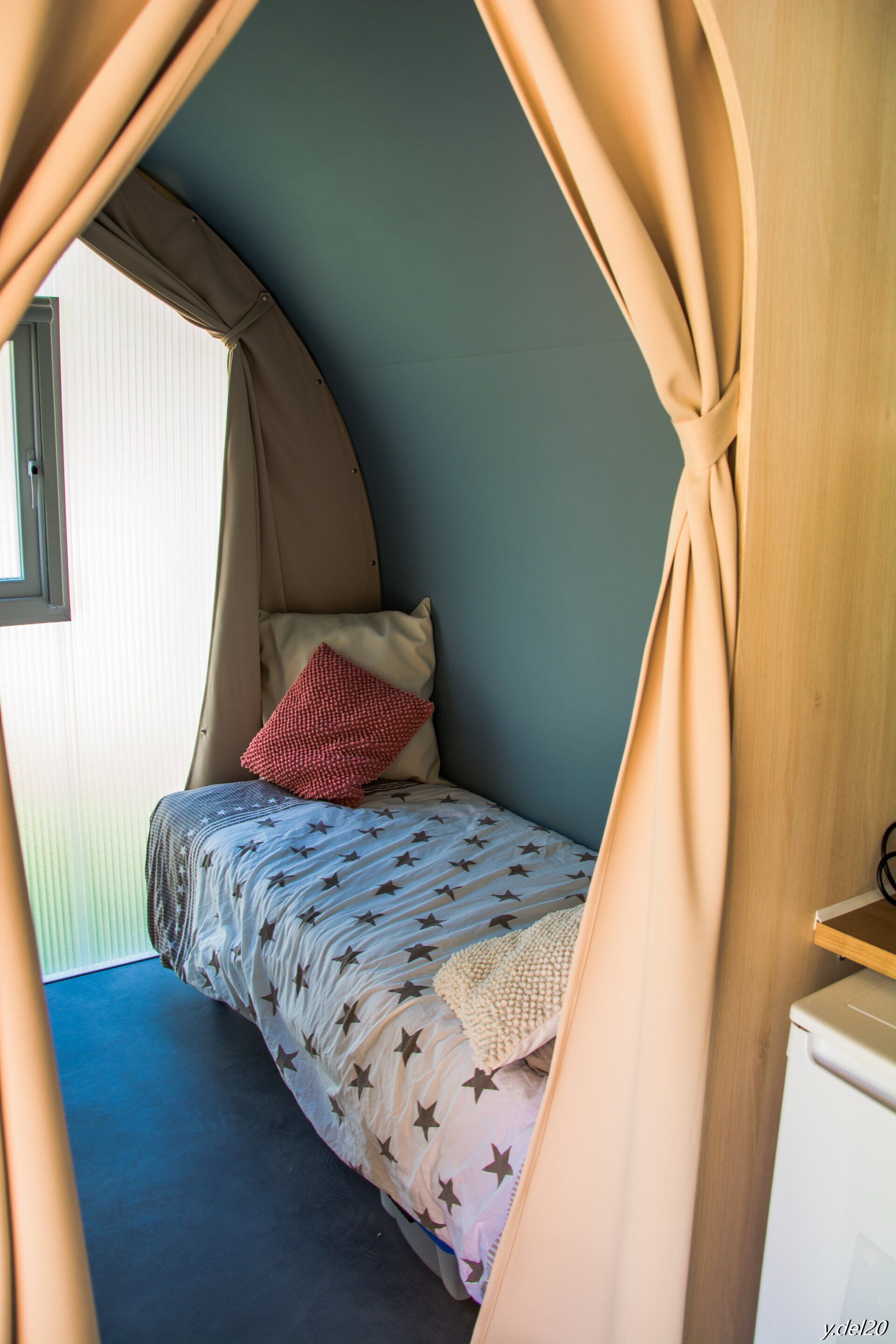 A cozy bedroom with a curved ceiling, a bed with star-patterned bedding, a red pillow, and a window on the left.