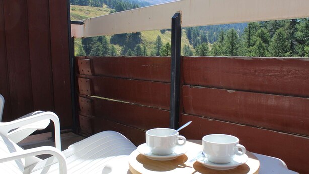 A balcony with a wooden railing, white plastic chairs, and a table with two white cups and saucers.
