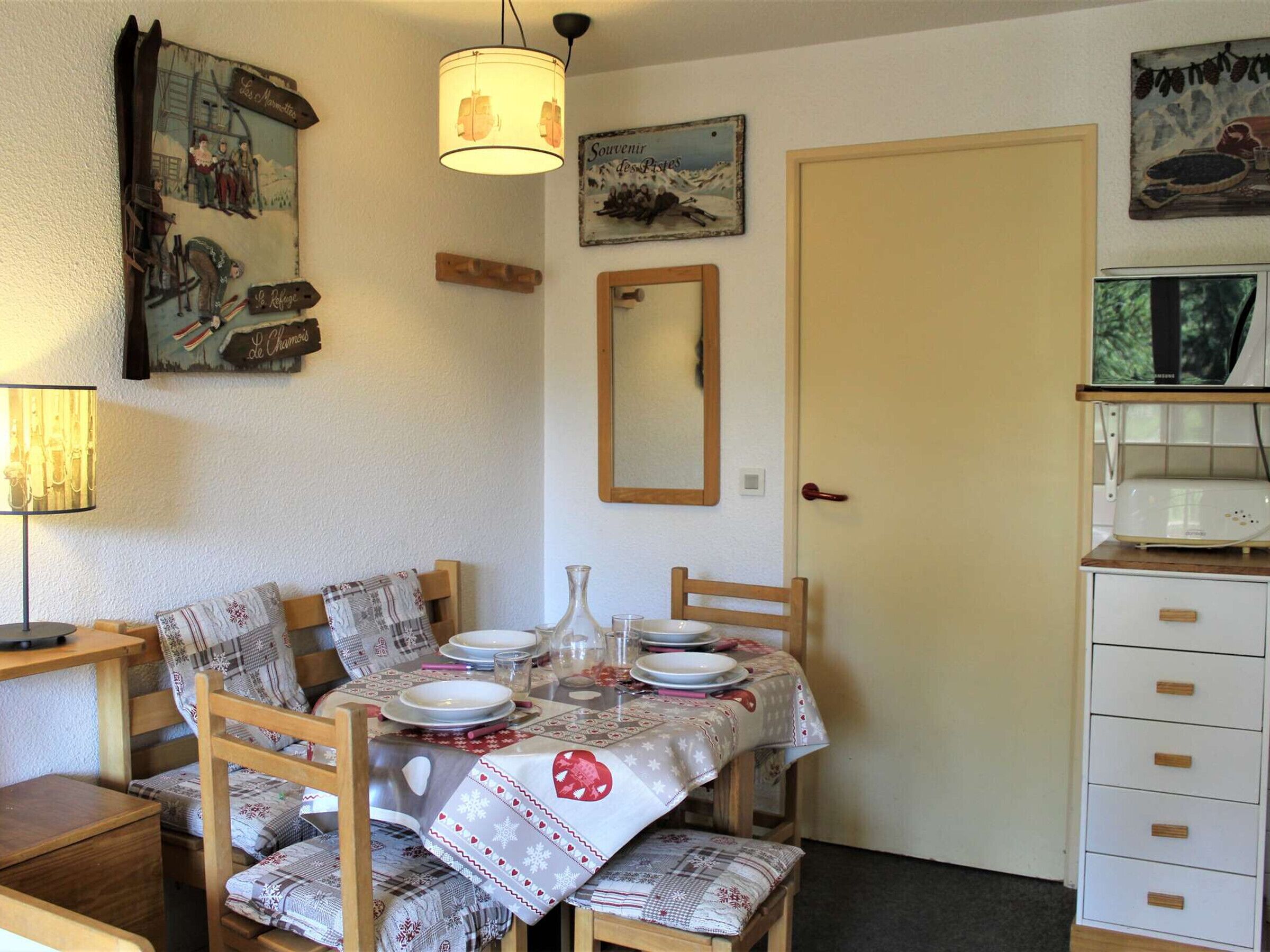 A dining area with a table set for four, wooden chairs, a wall-mounted mirror, and a shelf with a microwave.