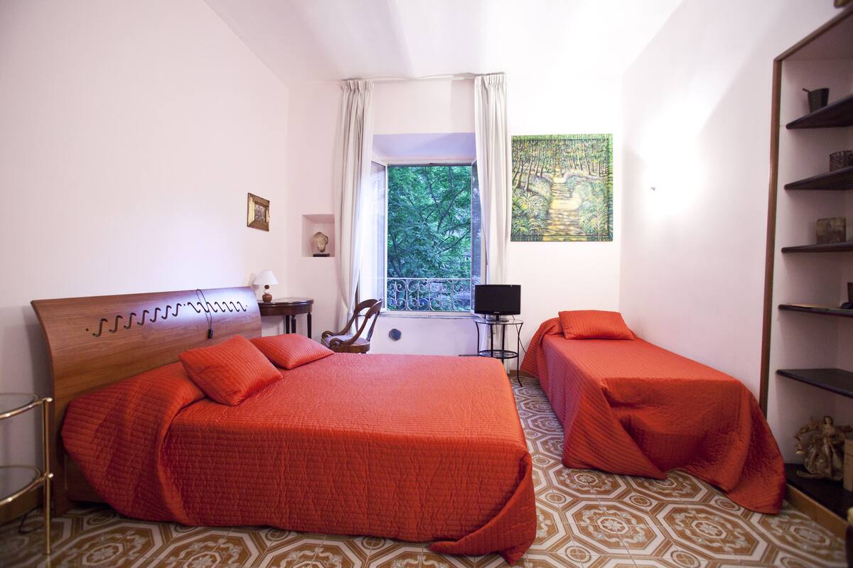 A bedroom with two beds, a window with a view of trees, a bookshelf, and a patterned floor.