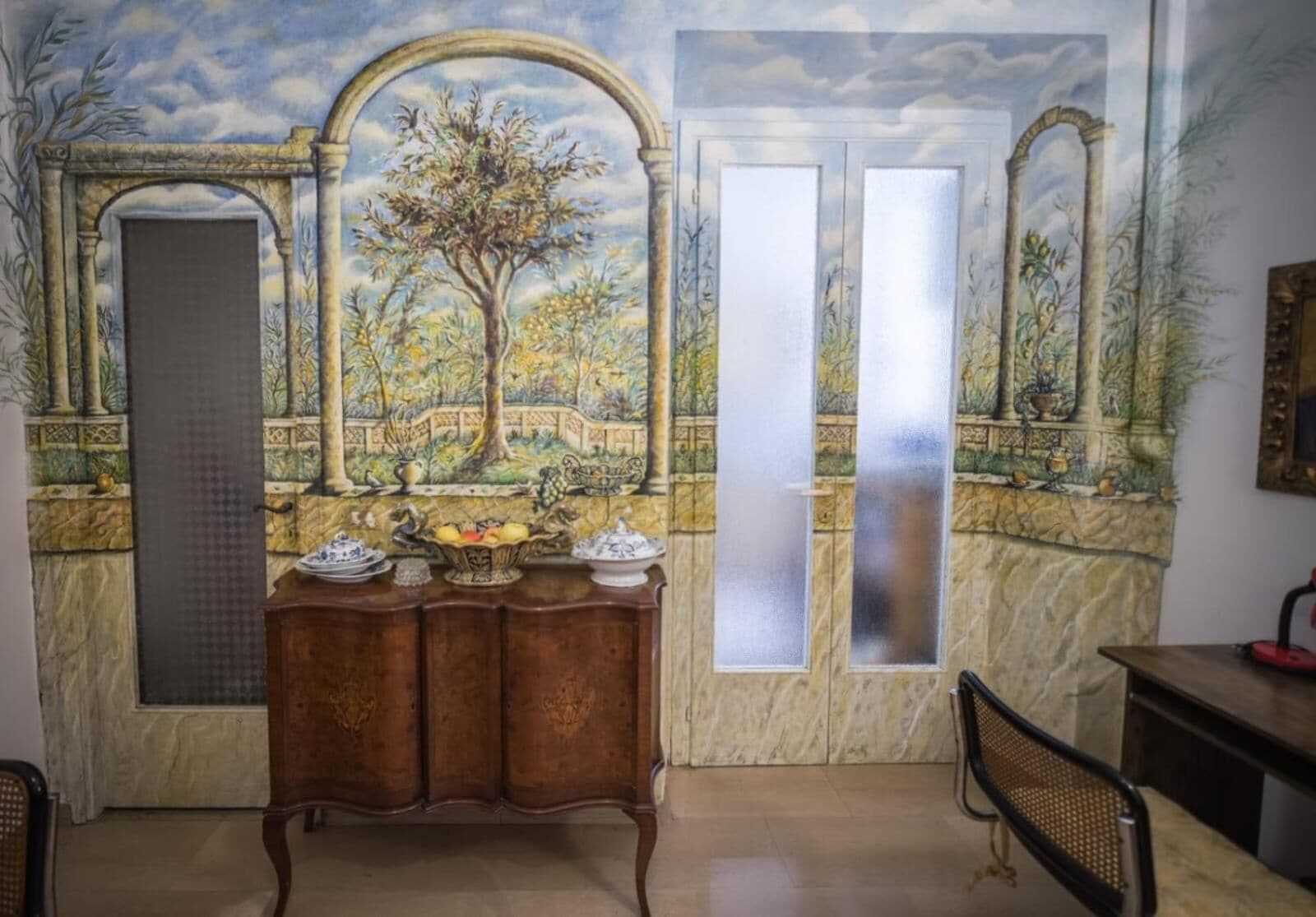 A dining area with a marble countertop, a wooden cabinet with dishes, and a mural of a garden scene.