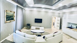 A modern living room with a sofa set, a round coffee table, a flat-screen TV mounted on the wall, and a kitchen area with cabinets and a sink. - Kreschatik Str two bedroom VIP (Shevchenkivs'kyi district)