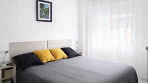 Un dormitorio con cama, mesita de noche, lámpara, un cuadro enmarcado en la pared y cortinas traslúcidas en la ventana.