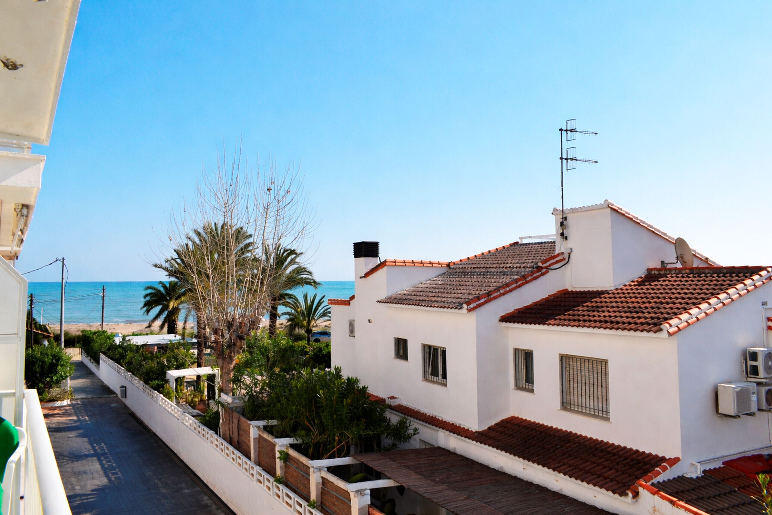 A coastal residential area with a white building featuring a tiled roof, a balcony, and a view of the sea.